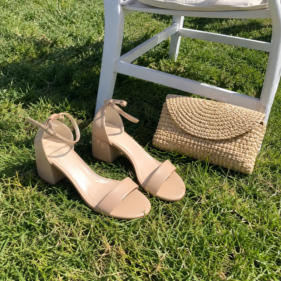 Nude block-heel sandals and a straw clutch on green grass beside a white chair